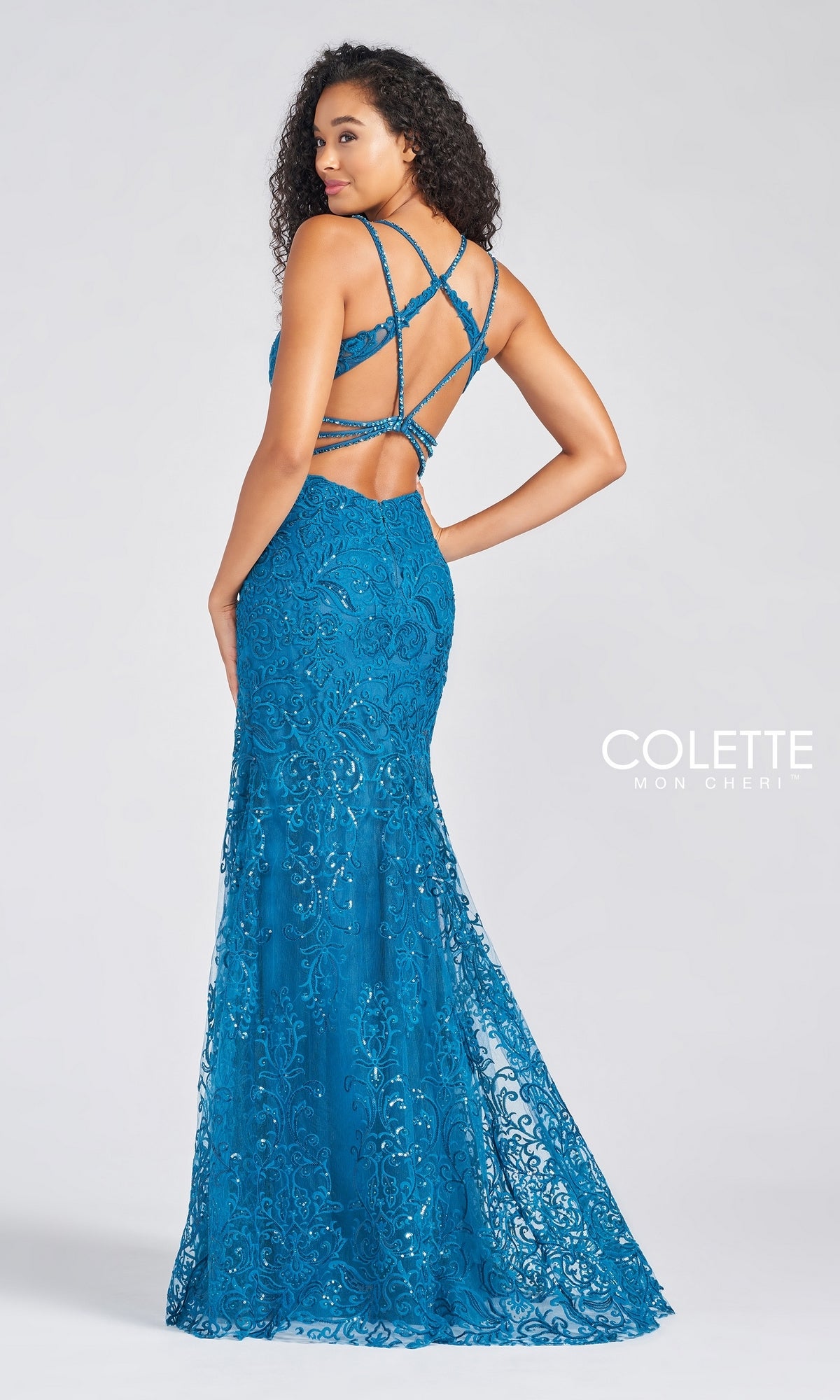 Woman wearing the Lace & Sequin Colette by Daphne Prom Dress CL12280, featuring blue embroidered lace, a fitted silhouette, and a crisscross strappy open back, stands looking over her shoulder.